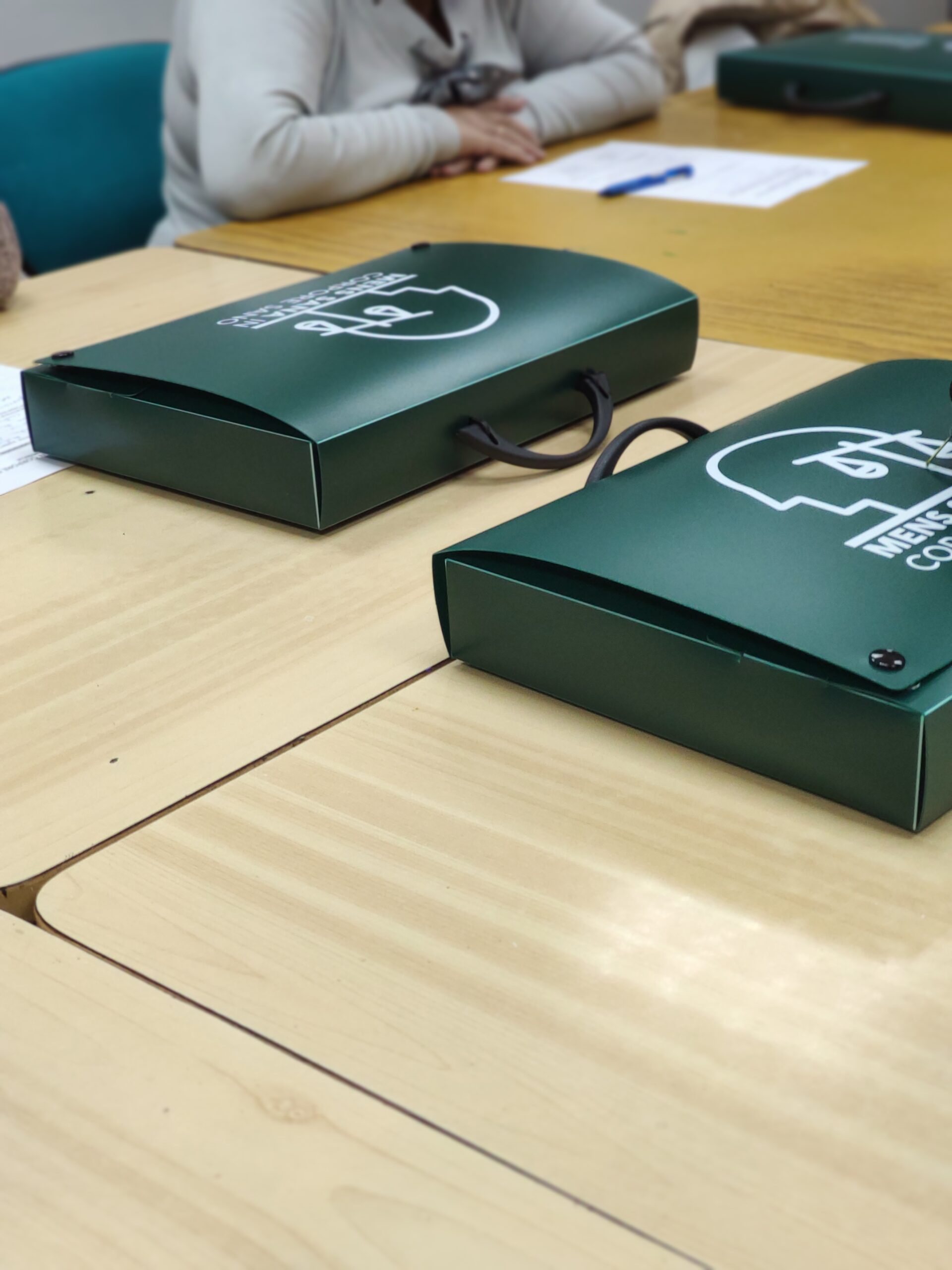 fotografía de una mesa de madera y encima de ella, posado el maletín rectangular y verde de formación UDP "Mens Sana in Copore Sano"